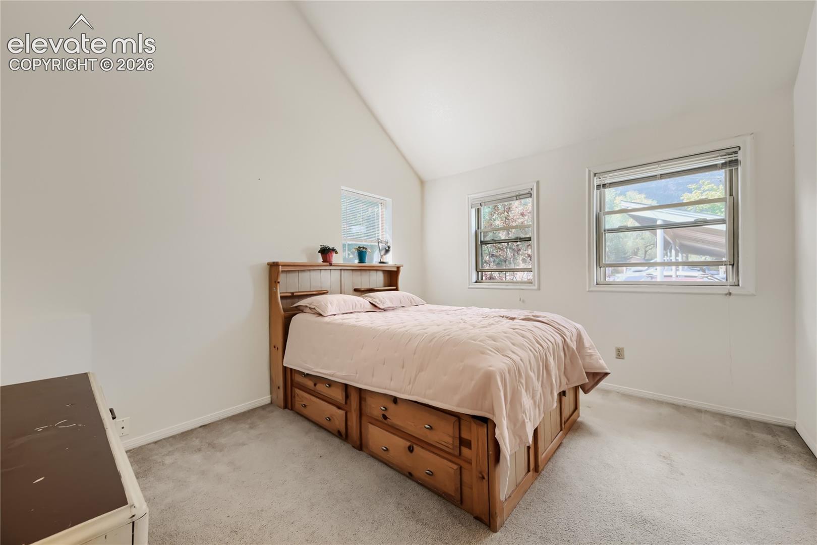 Bright secondary bedroom featuring vaulted ceilings, multiple windows with natural light, wood-finish bed frame with storage drawers, carpet flooring, and a compact dresser with mirror