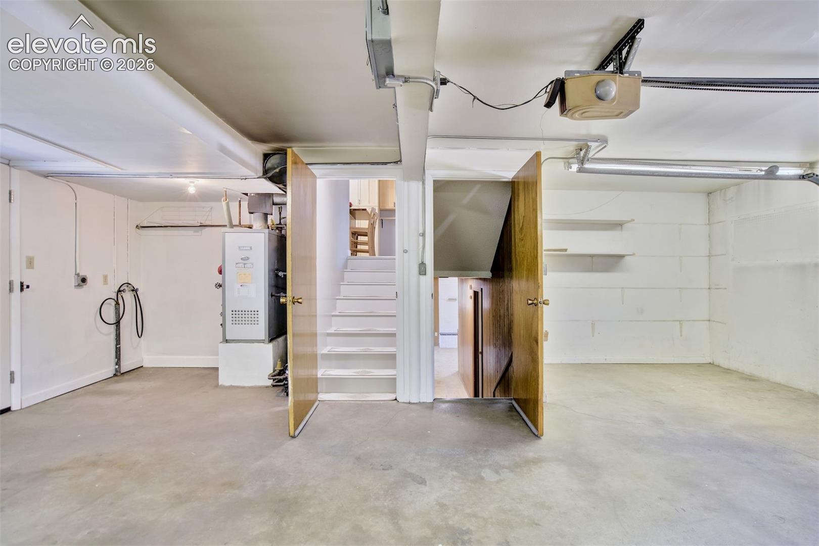 Enclosed garage space featuring painted masonry walls, concrete flooring, built-in shelving, and direct access to an interior staircase
