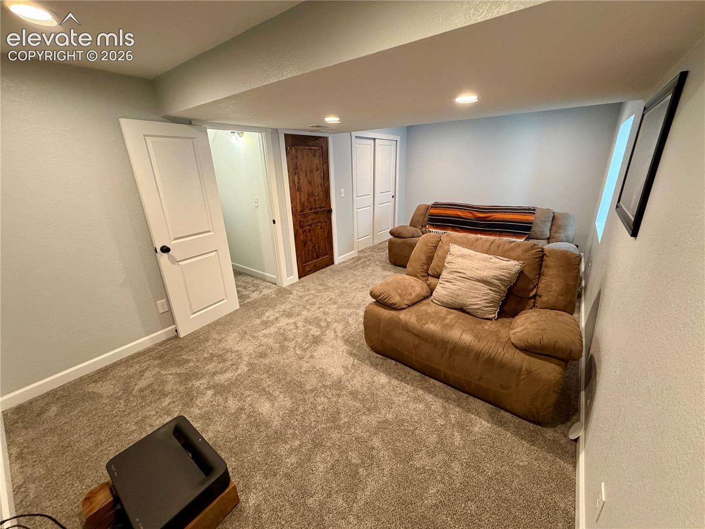 Basement carpeted room (bedroom or media room) featuring light gray walls, recessed lighting, white baseboards, a white two-panel door with black hardware, and a dark wood-finish two-panel door