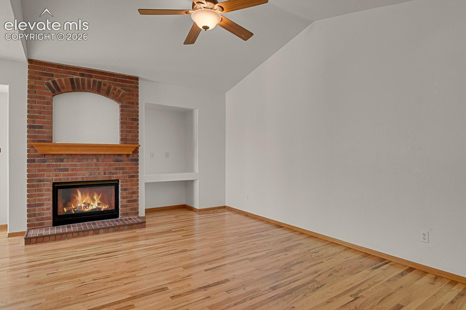 Brick fireplace with wood mantel, built-in shelving, vaulted ceiling, wood-finish flooring, and ceiling fan with light fixture