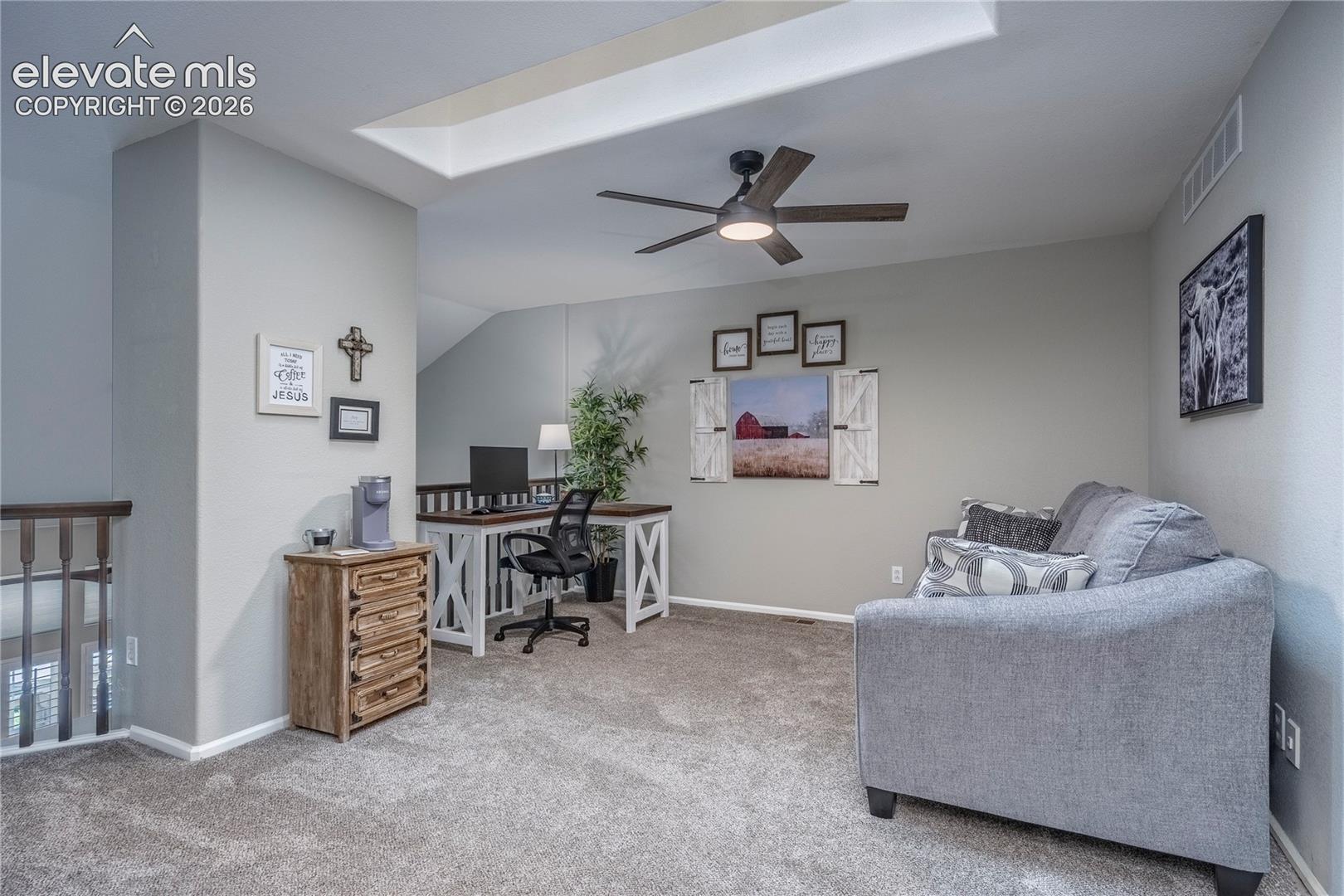 Open-concept room featuring a skylight, ceiling fan with integrated lighting, neutral wall-to-wall carpeting, and a wooden stair railing with spindles