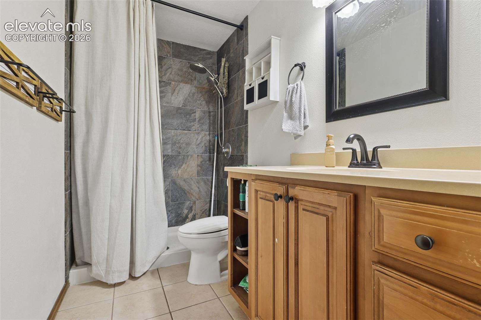 Bathroom featuring a tiled shower surround, a vanity with a light-toned countertop, wood-finish cabinetry, and a dark-framed mirror