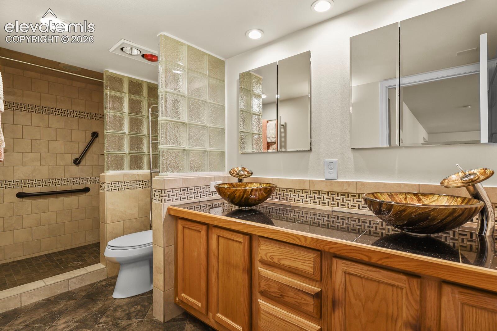 Dual wood-finish vanity with amber-toned glass vessel sinks, stone-tiled walk-in shower with integrated glass block partition, recessed lighting, and wall-mounted medicine cabinets