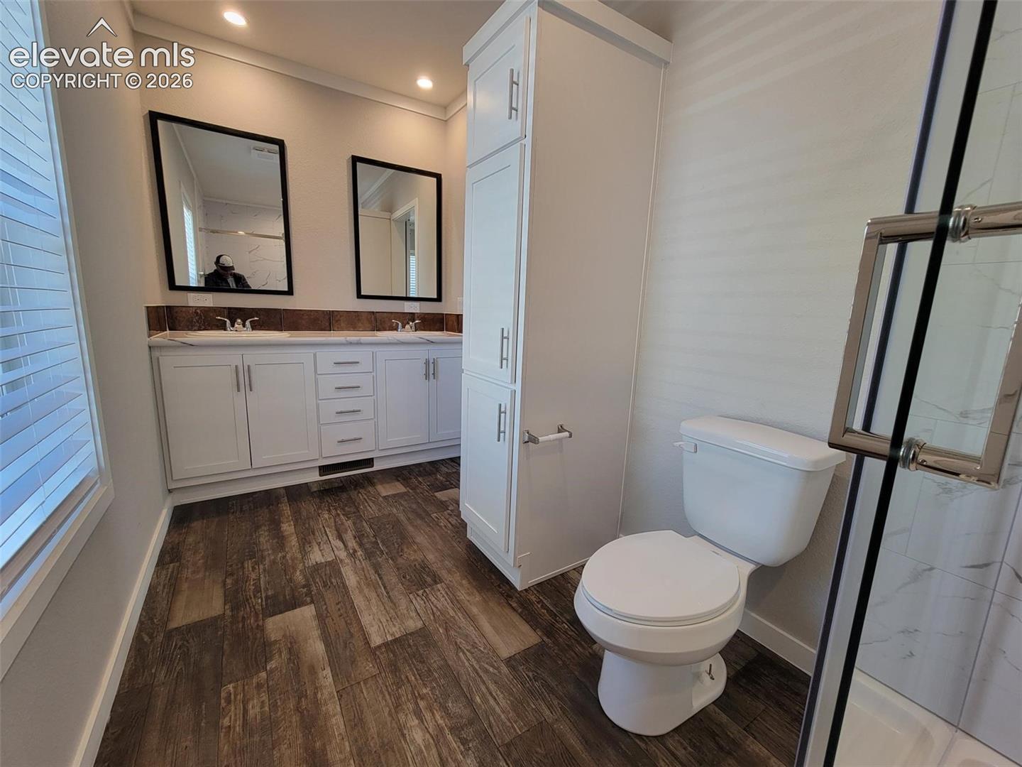 Bathroom featuring a dual-sink vanity with white cabinetry, wood-finish flooring, full-height linen storage, and a glass-enclosed shower with tiled surround