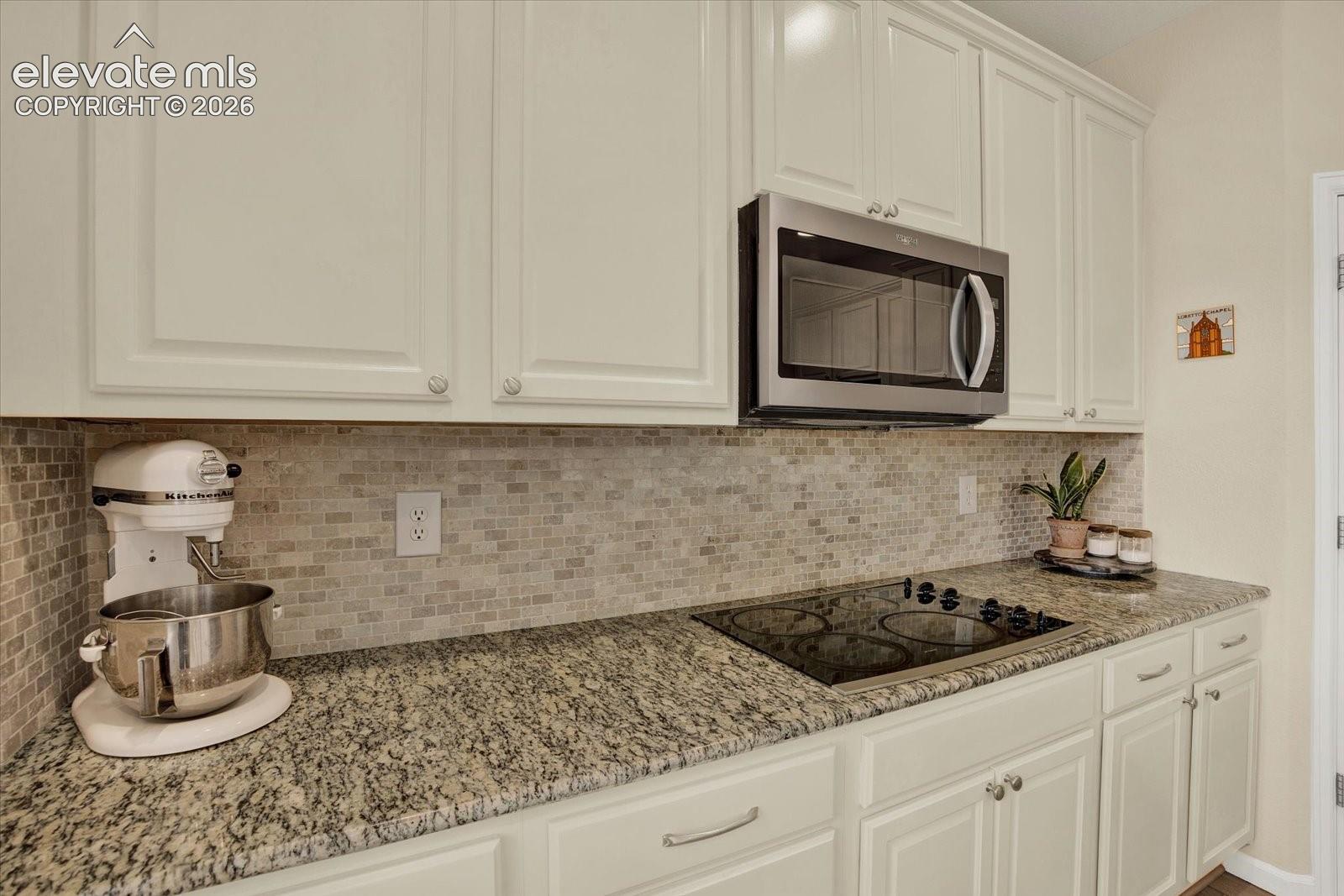 Off-white cabinetry, stone-patterned countertops, mosaic tile backsplash, built-in stainless steel microwave, and integrated cooktop