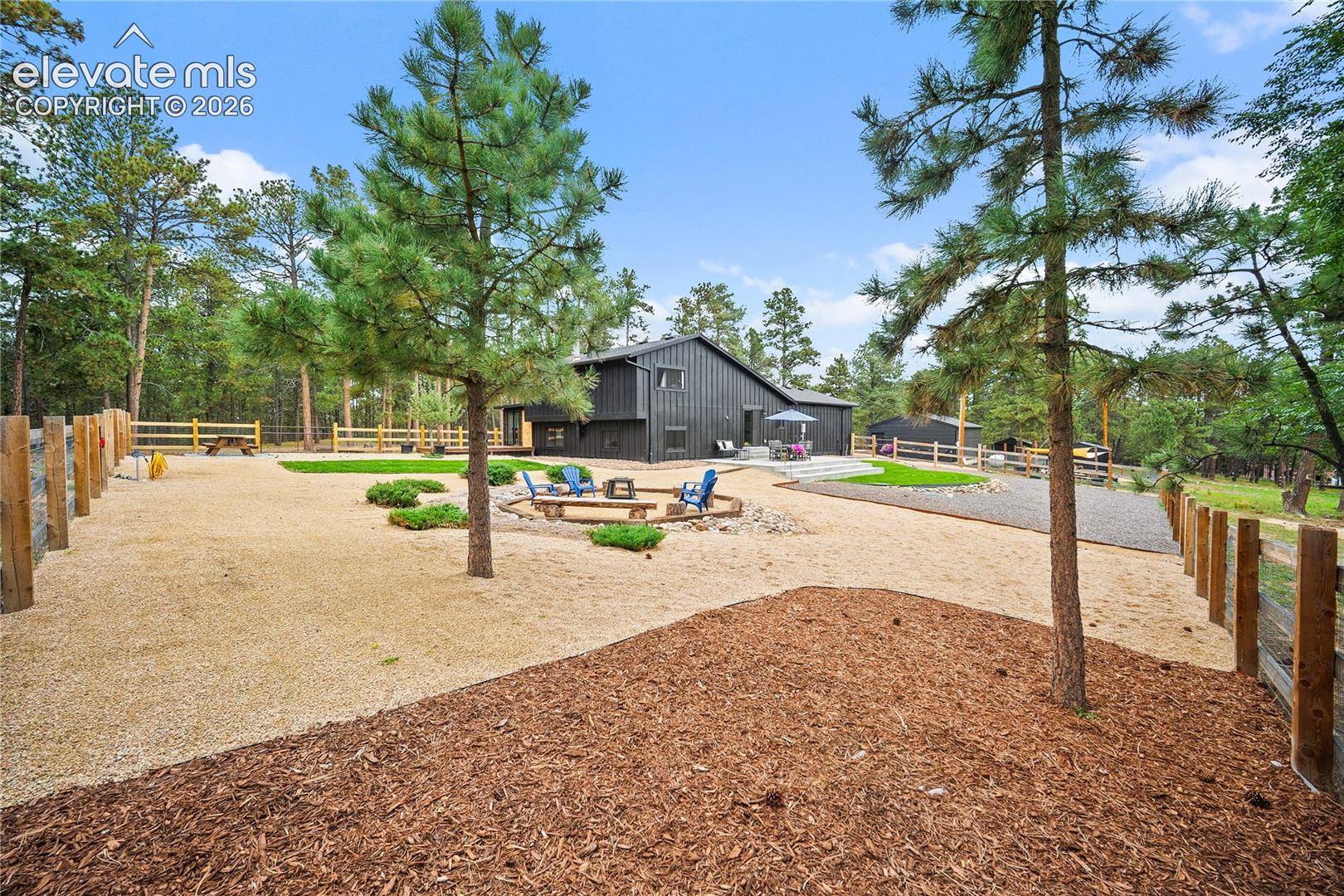 Expansive backyard featuring tiered wood fencing, gravel and mulch ground cover, established pine trees, and a stone-bordered fire pit area