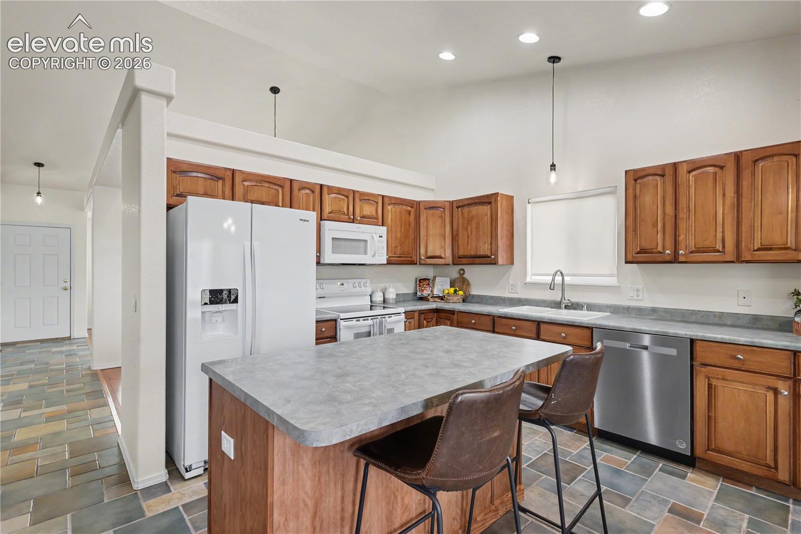 Expansive kitchen featuring a central island with seating, wood cabinetry, stainless steel dishwasher, white appliances, and a vaulted ceiling