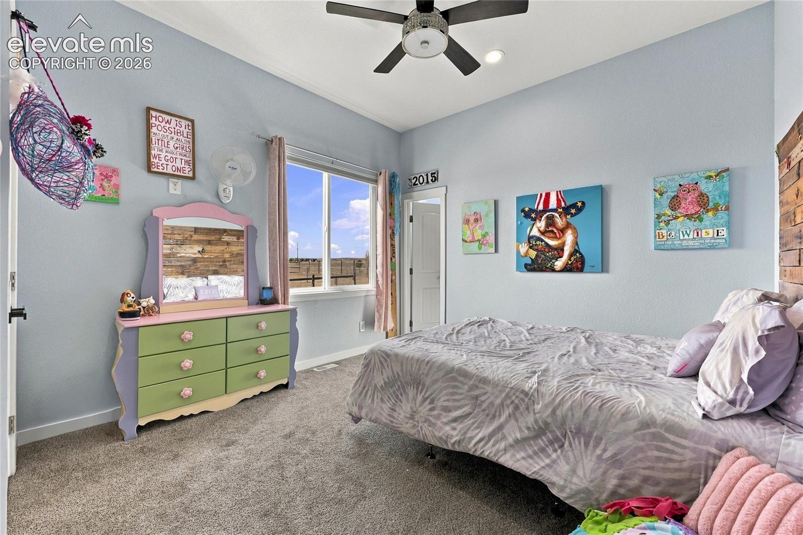 Neutral-toned bedroom featuring carpeted flooring, vaulted ceilings with an integrated multi-blade ceiling fan, and a single-pane window with exterior landscape exposure