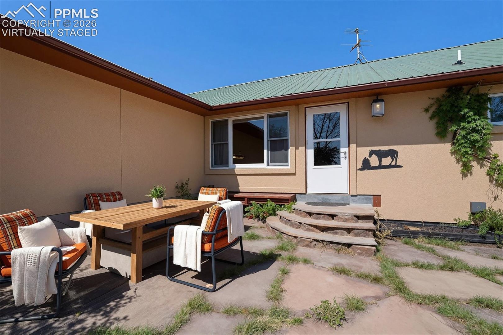 virtually staged Outdoor patio area featuring large flagstone pavers, a light tan stucco exterior, a green metal roof, and a white entry door with an exterior light fixture