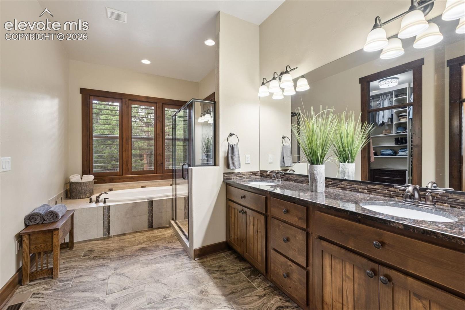 Dual-sink vanity with stone countertops and wood cabinetry, glass-enclosed shower, built-in soaking tub, and tile flooring