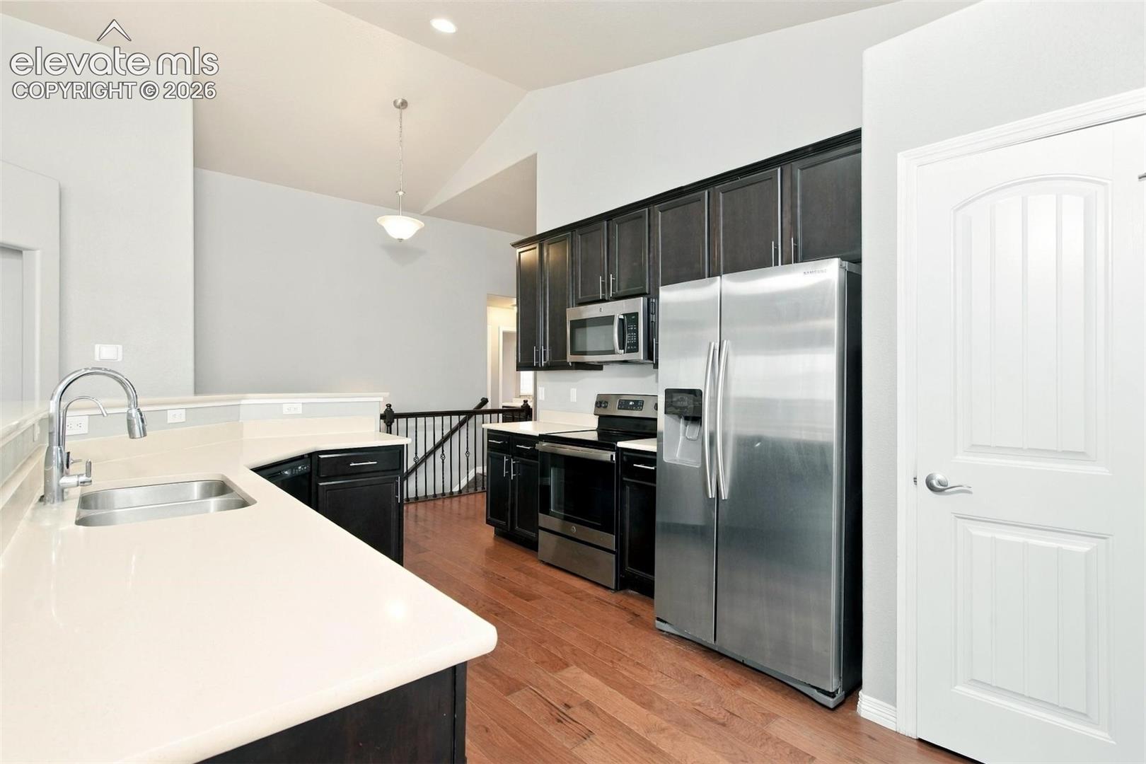 Kitchen featuring dark wood cabinetry, solid-surface countertops, stainless steel appliances, and wood-finish flooring under a vaulted ceiling