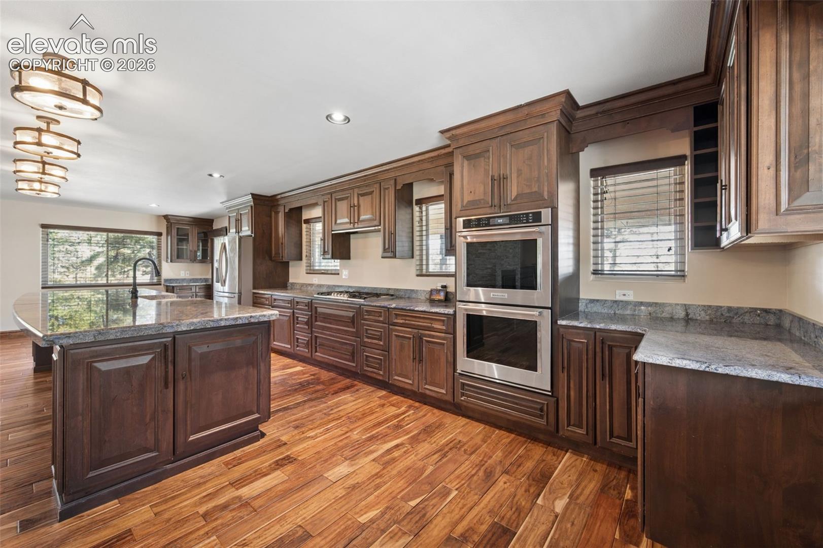 Kitchen featuring wood cabinetry, natural stone countertops, a multi-level center island, wood-finish flooring, and stainless steel double wall ovens