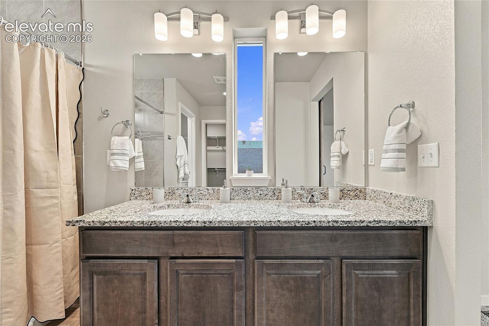 Dual vanity with integrated sinks, granite countertop, rich wood-finish cabinetry, and overhead sconce lighting