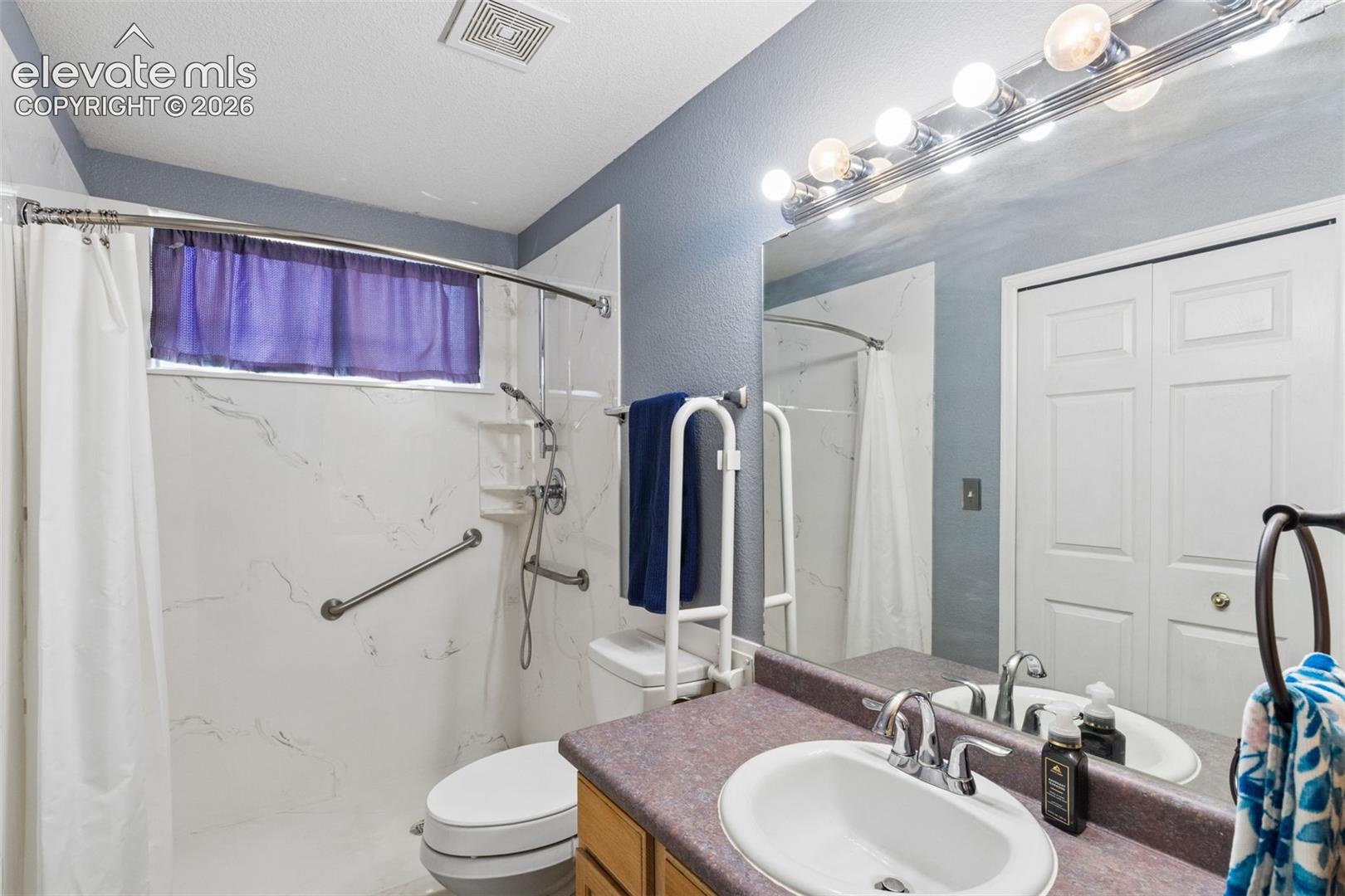 Bathroom featuring a solid-surface vanity countertop, undermount sink with dual-handle faucet, full-width vanity mirror with integrated lighting, and a shower enclosure with floor-to-ceiling stone-patterned wall surround
