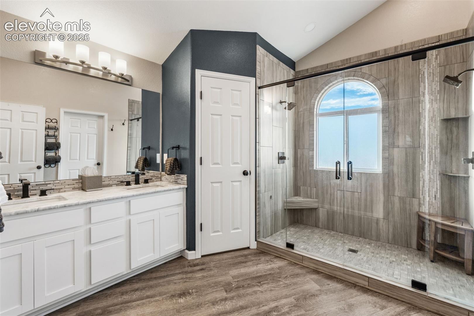 Dual-vanity cabinet with stone-finish countertop, walk-in shower with floor-to-ceiling tile, arched window, built-in shower seating, and wood-finish flooring