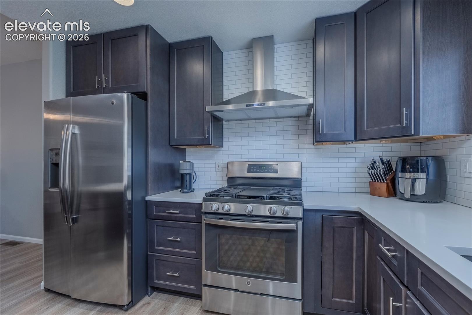 Kitchen featuring dark-finish cabinetry, stainless steel appliances, quartz countertops, white subway tile backsplash, and wood-finish flooring