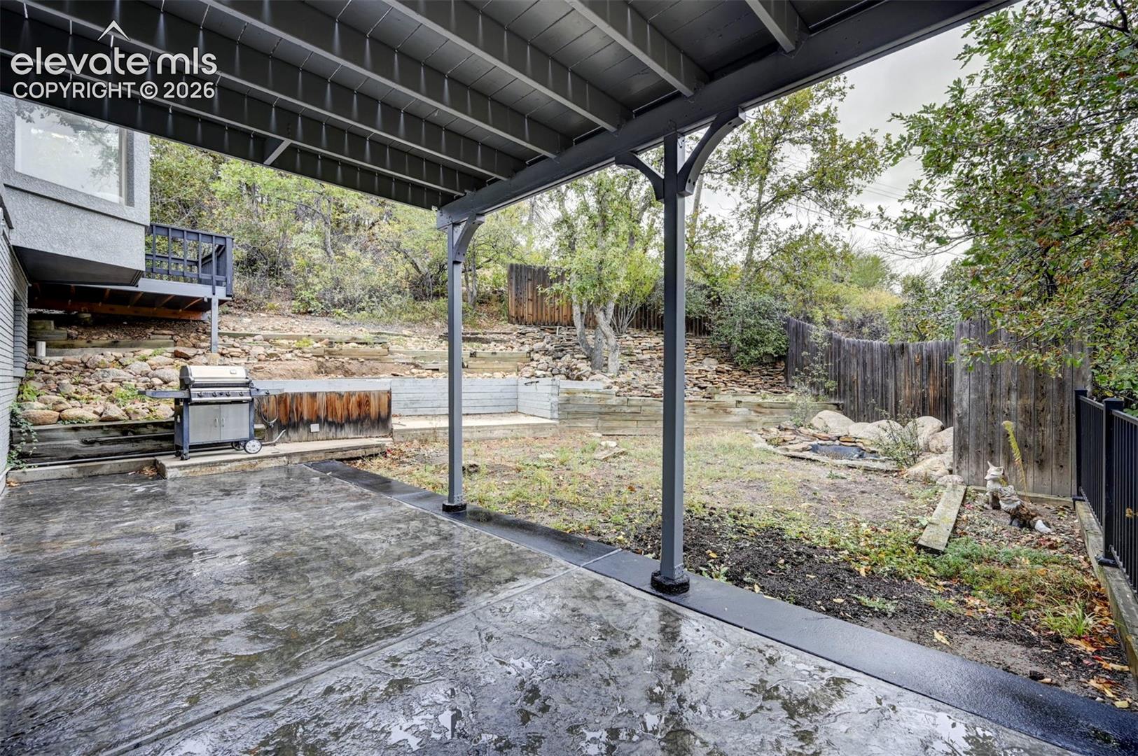 Covered concrete patio featuring structural support columns, tiered stone retaining walls, and wood fencing