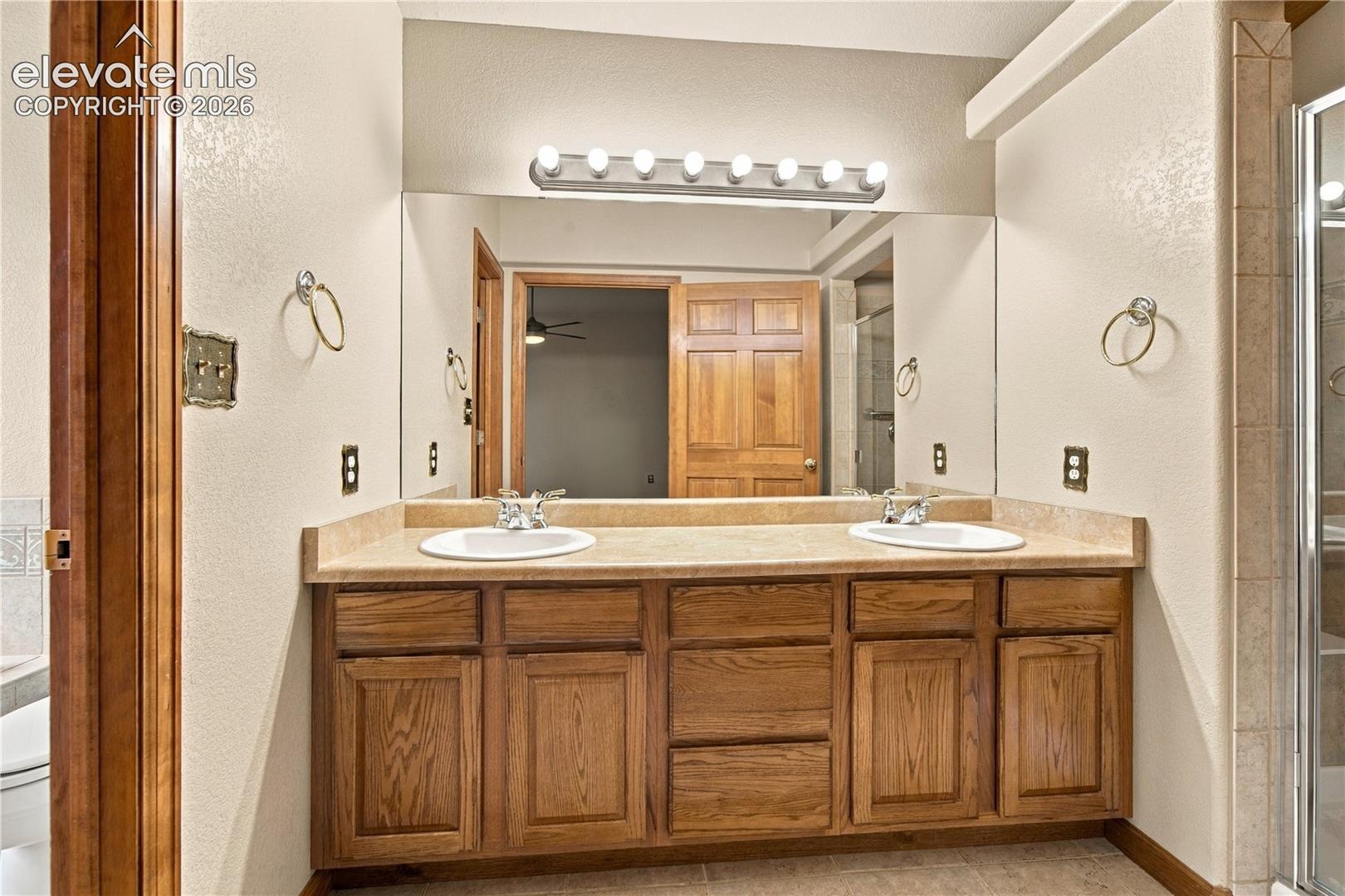 Double vanity featuring a light-toned stone countertop, dual undermount sinks, solid wood cabinetry, a full-width wall mirror, and an eight-bulb vanity light fixture