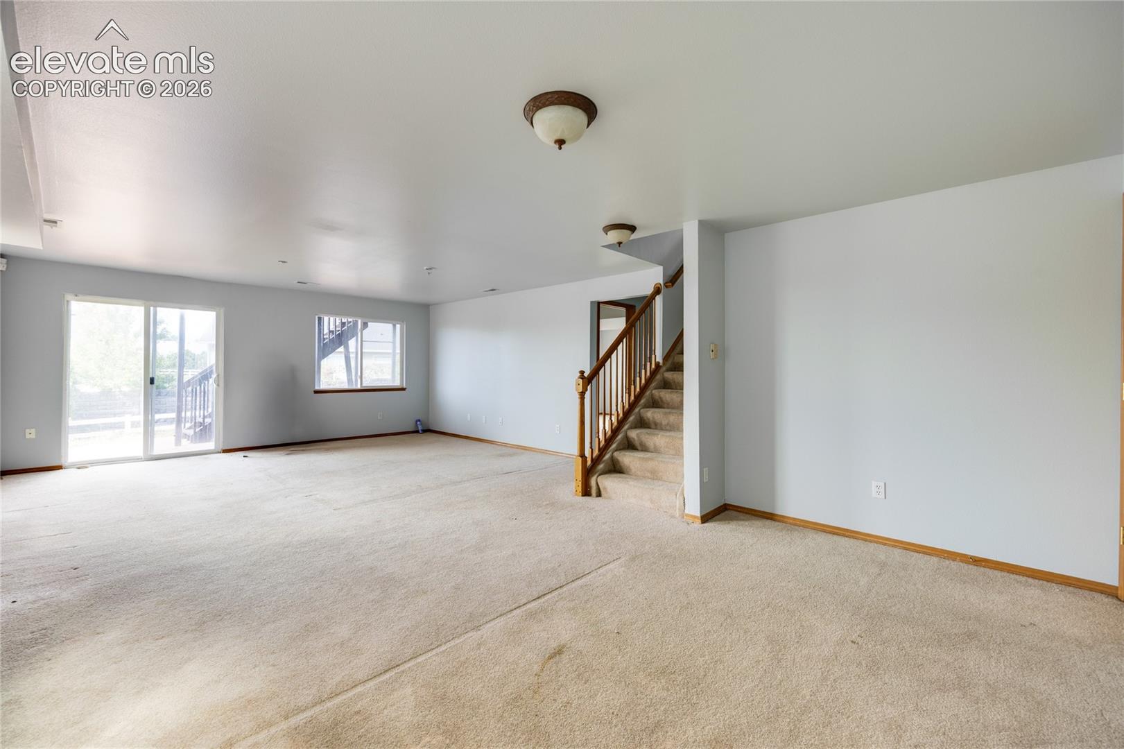 Spacious room featuring carpeting, a sliding glass door, a window, a wooden staircase with carpeted treads, and ceiling-mounted light fixtures