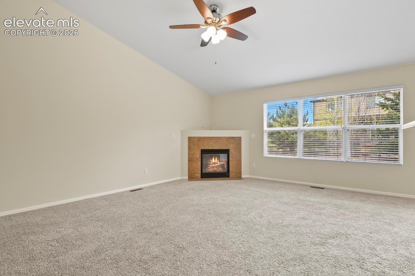Spacious living area featuring a vaulted ceiling with a ceiling fan, a gas fireplace with a tiled surround, and large windows providing abundant natural light