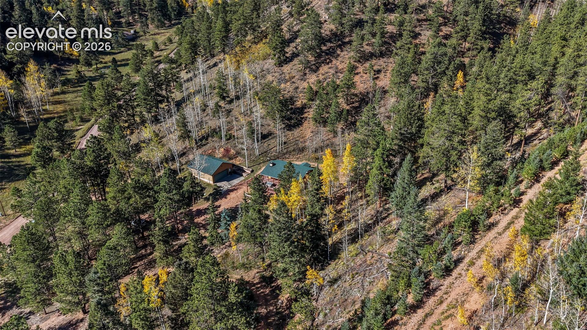 Aerial perspective of a multi-structure property featuring metal roof profiles, exterior wood siding, and surrounding dense evergreen and deciduous forest landscape