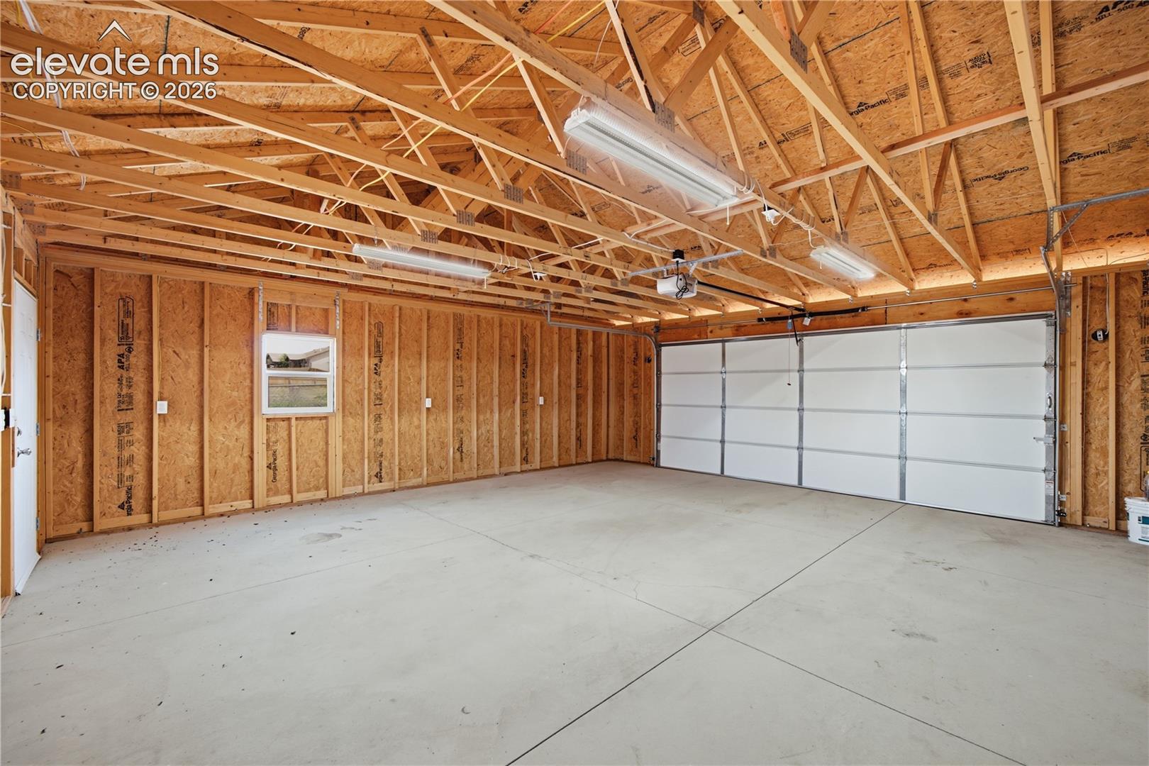 Spacious garage featuring exposed wood-beam ceiling construction, polished concrete flooring, white paneled overhead door, and wood-paneled walls with integrated storage cabinetry