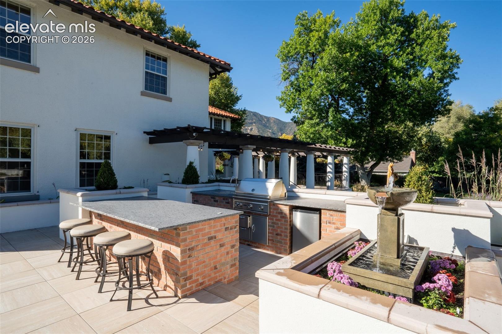 Outdoor kitchen featuring brick masonry, stone countertops, a stainless steel grill, and an integrated beverage refrigerator, complemented by a stone-tiled patio, a tiered water fountain, and a column-supported pergola
