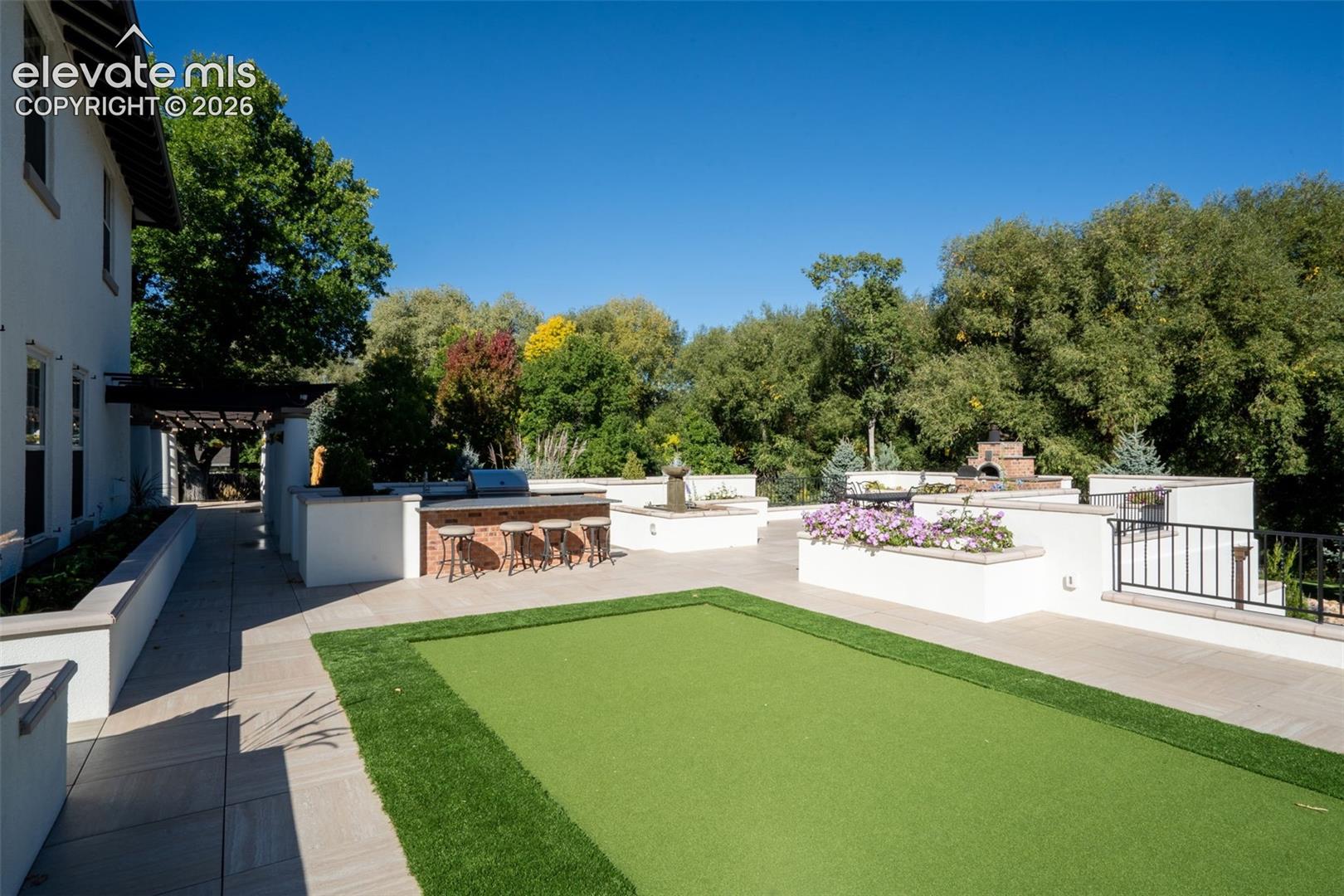 Expansive stone-paved terrace featuring a built-in outdoor kitchen with brick detailing, integrated artificial turf putting green, elevated stone planters, and decorative wrought-iron railings