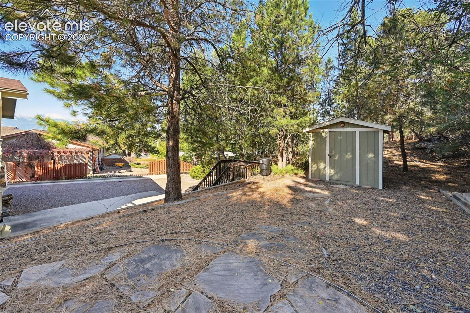 Exterior grounds featuring a stone-paver patio, detached storage shed, mature coniferous trees, and wood-railing stairs