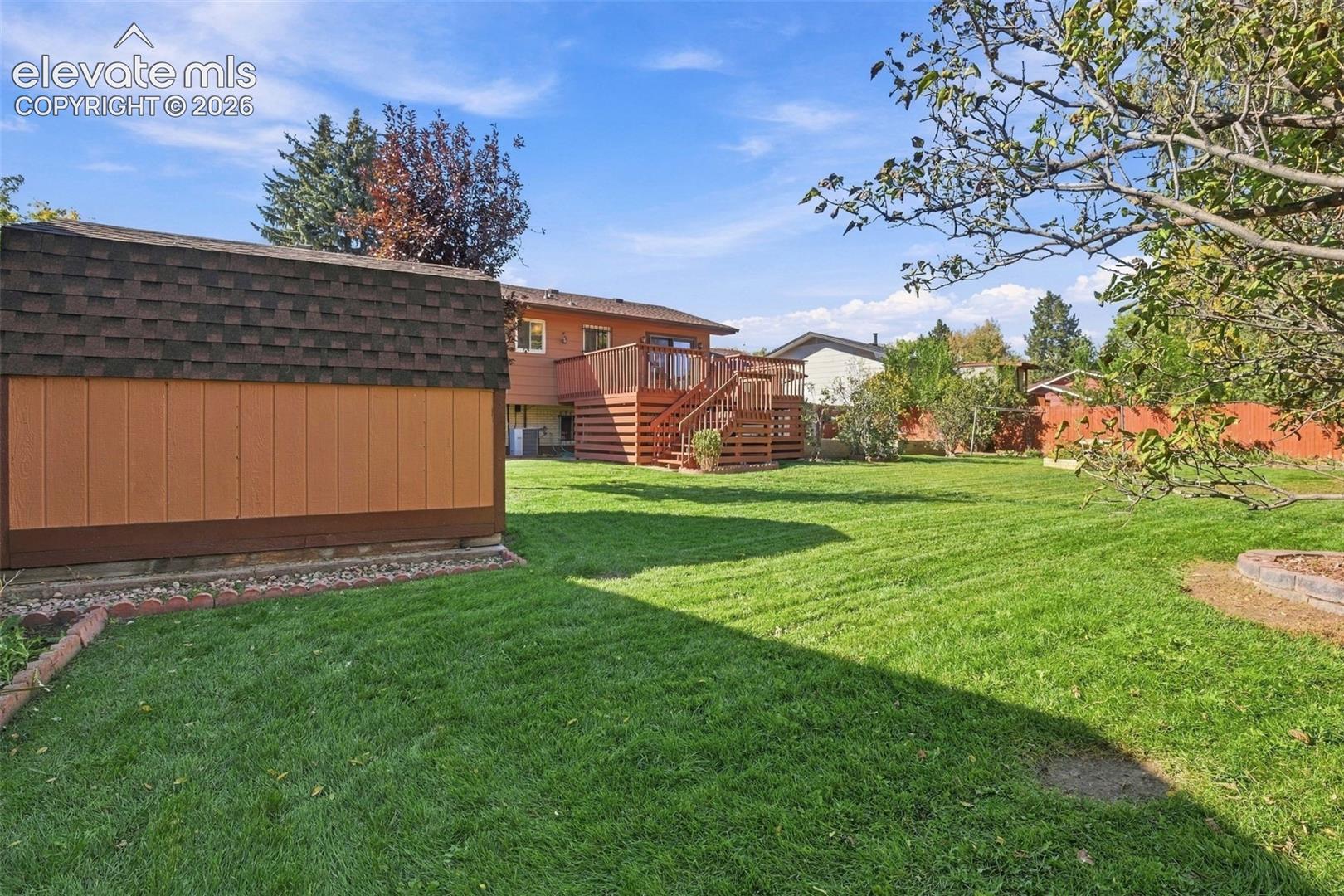 Expansive backyard featuring a manicured lawn, detached storage structure with shingle roofing, elevated wood deck with stairs, and stone-bordered landscaping beds