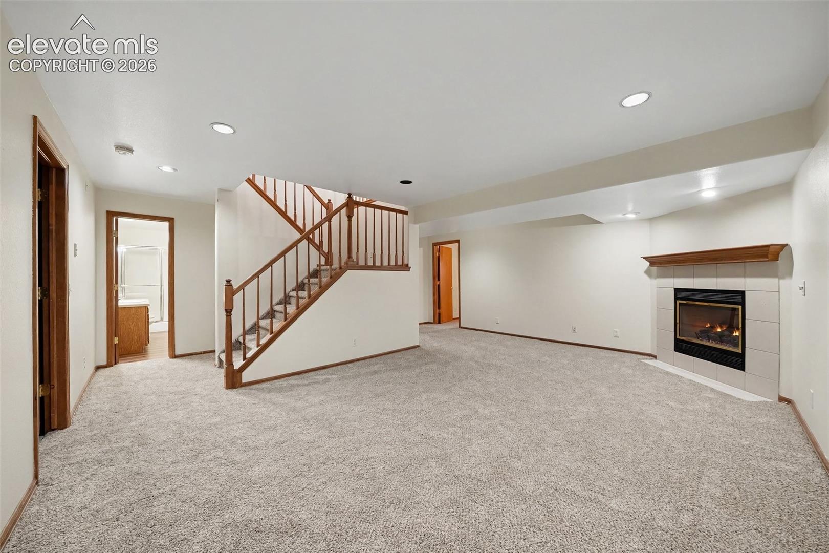 Spacious room featuring light gray carpet, recessed lighting, a fireplace with a tiled surround, and a wood-finish staircase