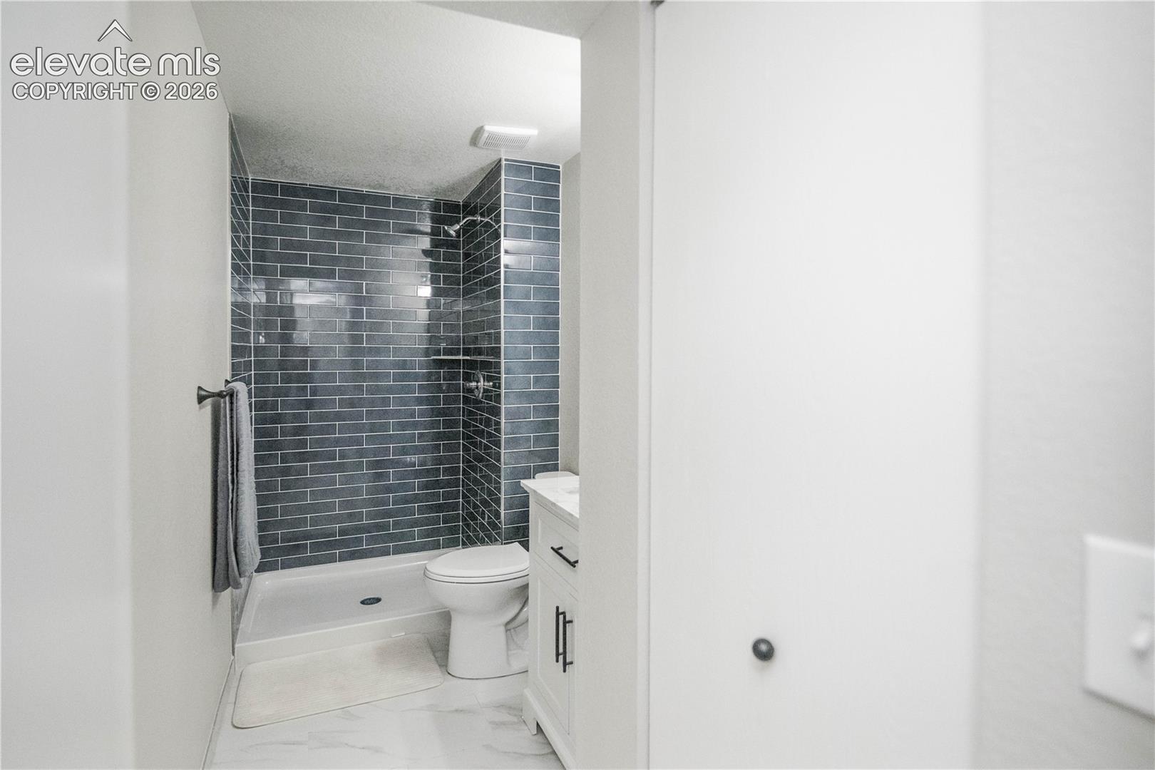 Bathroom featuring a walk-in shower with blue subway tile surround, white vanity with integrated hardware, and stone-finish flooring