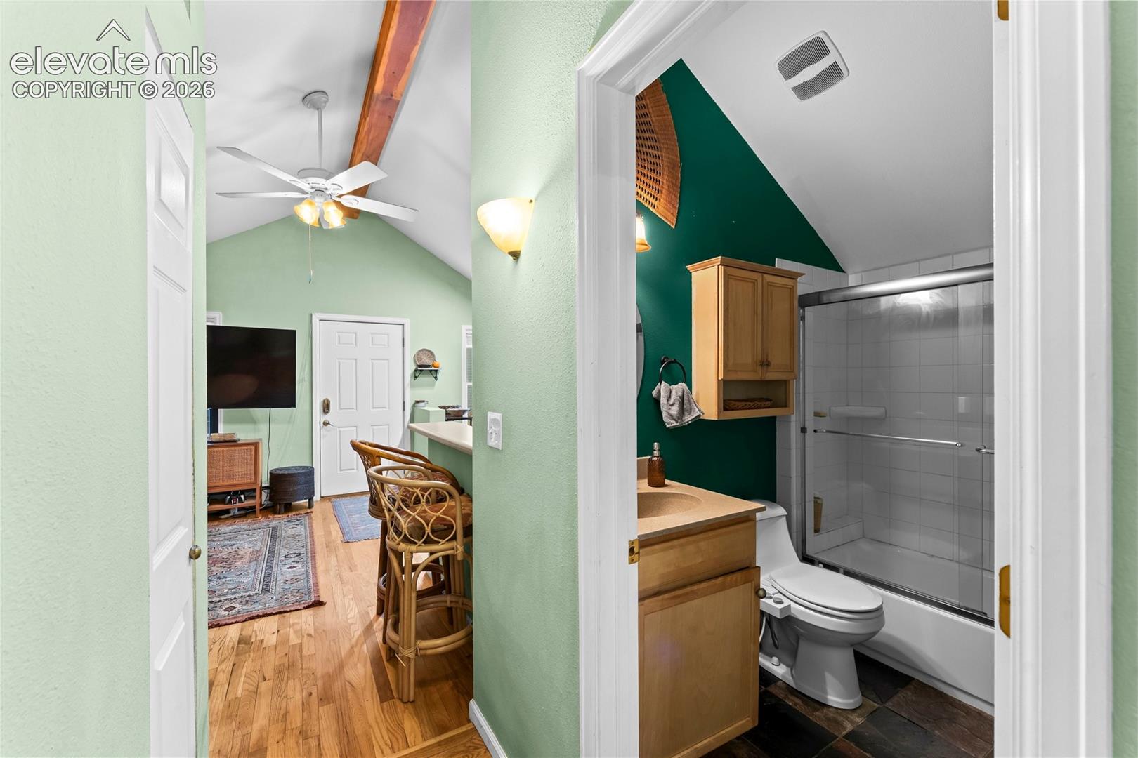Vaulted ceilings with exposed wood beam, ceiling fan, wood-finish flooring, and bathroom featuring a wooden vanity cabinet and a shower-tub combination with tile surround
