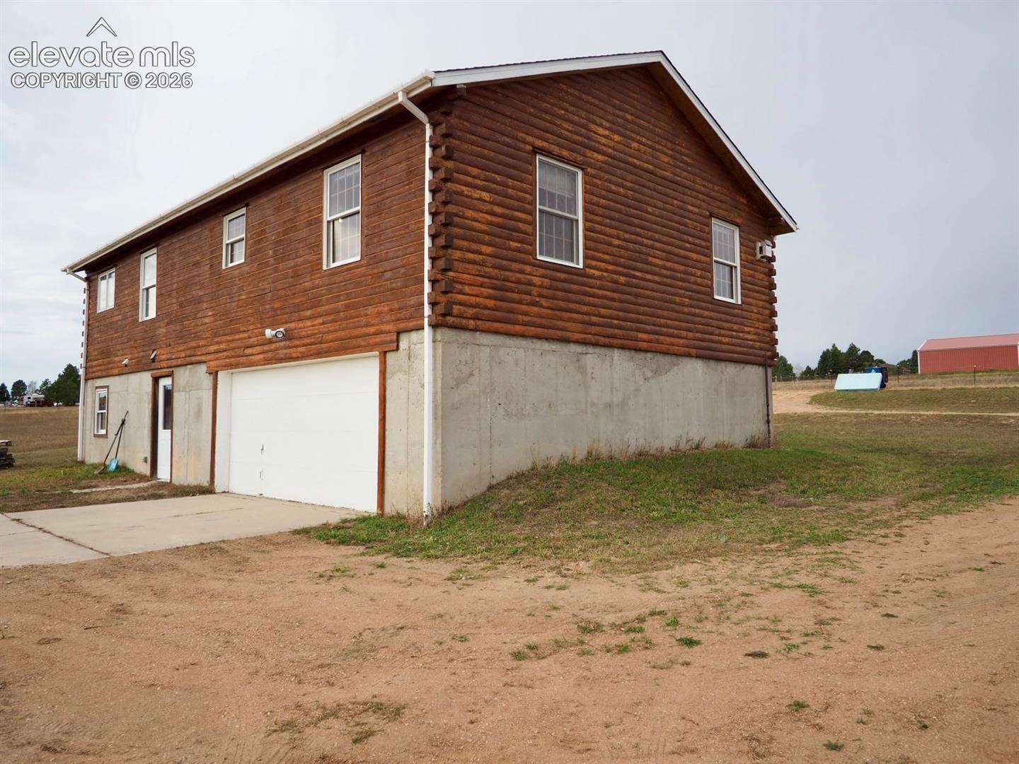 Two-story log cabin exterior featuring a concrete foundation, attached garage with white roll-up door, multiple double-hung windows, and a side entry door