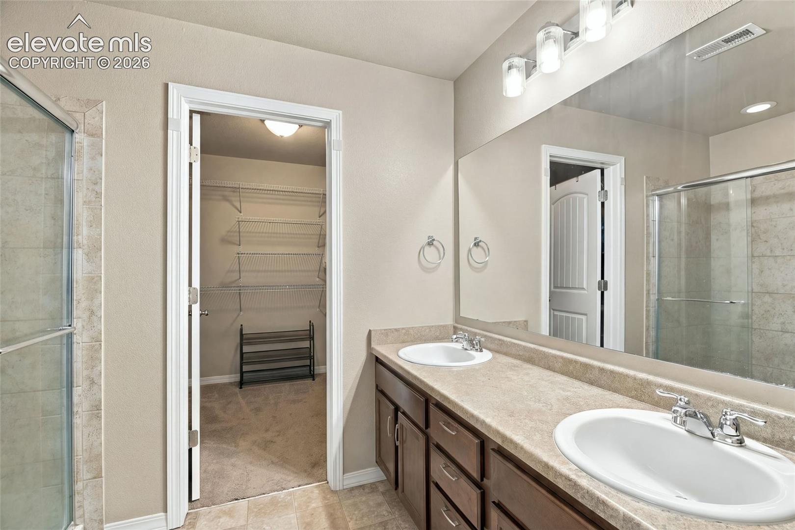 Dual-sink vanity with stone-finish countertop, full-width wall mirror, glass-enclosed shower with tile surround, and attached walk-in storage area with wire shelving
