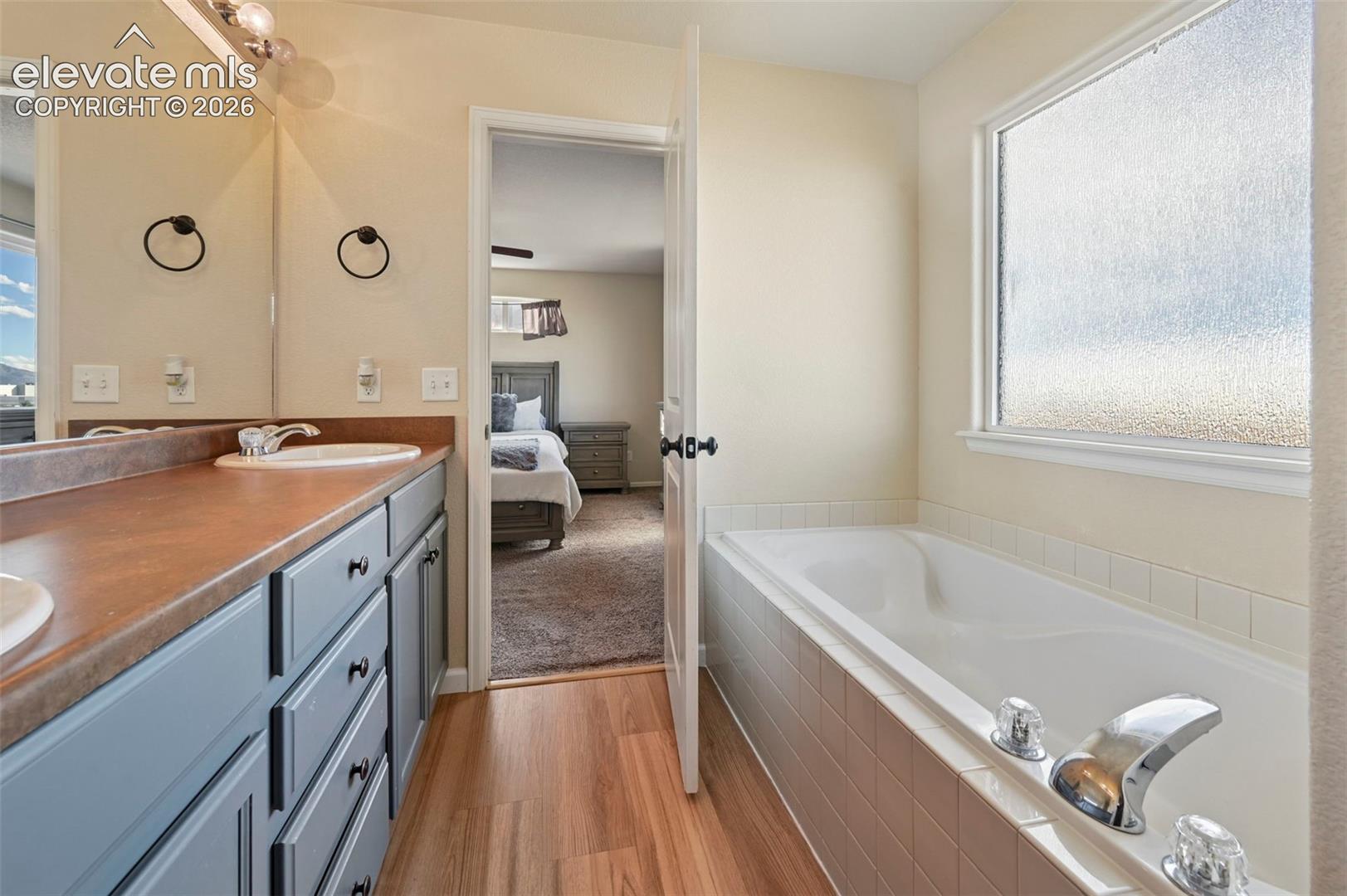 Bathroom featuring a dual-sink vanity with matte-finish cabinetry, wood-finish flooring, a soaking tub with tiled surround, and a wall-to-wall vanity mirror