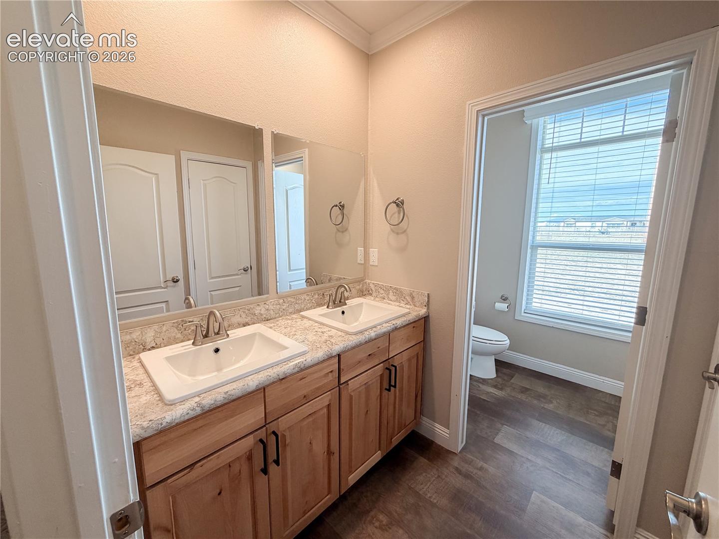 Dual-sink vanity with natural wood-finish cabinetry, stone-patterned countertops, drop-in rectangular basins, polished chrome fixtures, and wood-finish flooring