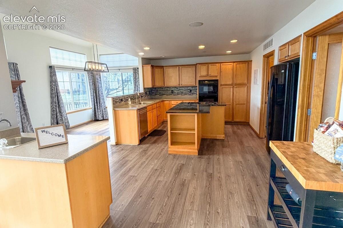 Open-concept kitchen with wood-finish flooring, light wood cabinetry, a central island with dark countertops, a built-in oven, and recessed lighting