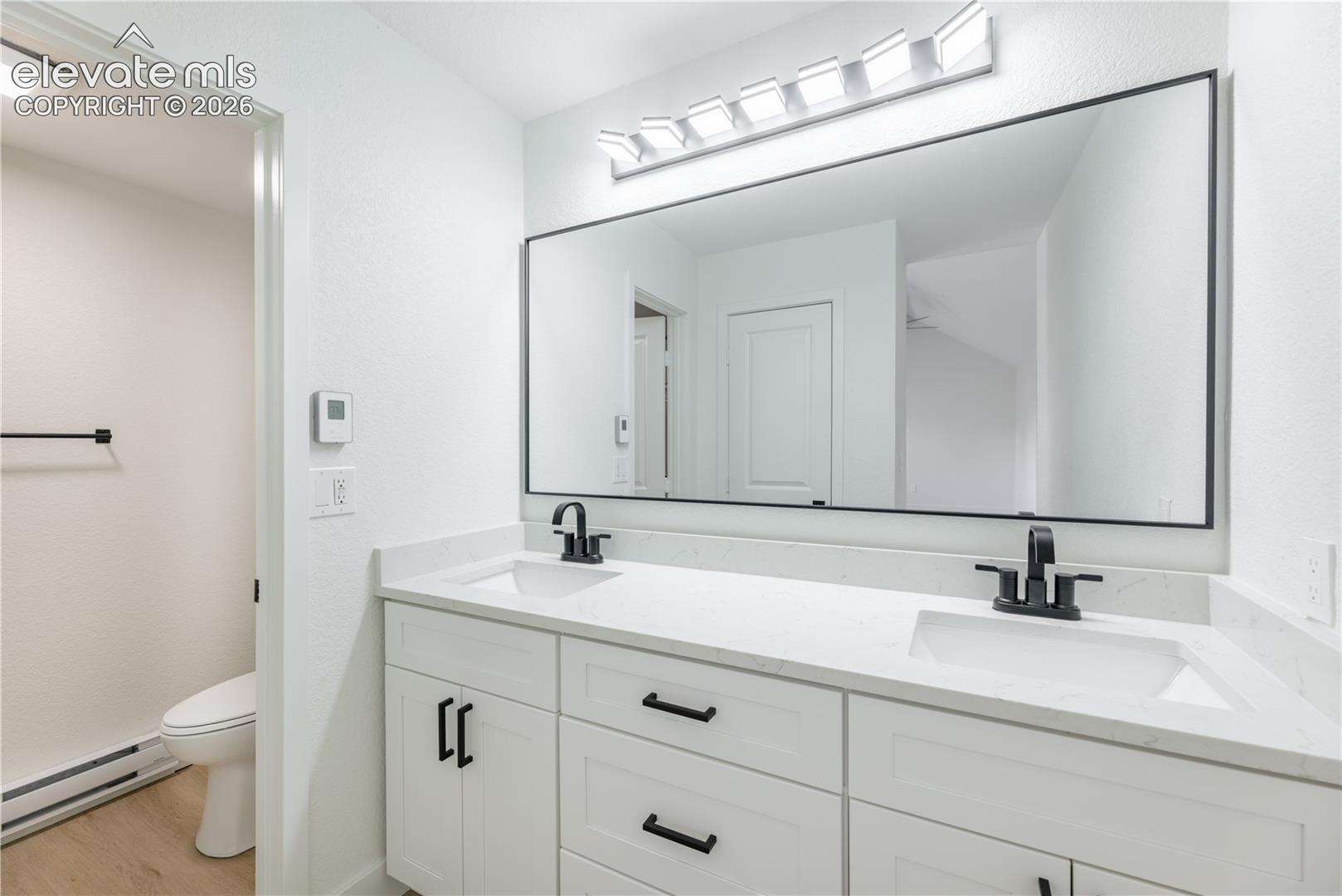 Dual-sink vanity featuring a stone countertop, white shaker-style cabinetry, matte black hardware, and an expansive wall-mounted mirror with integrated multi-light fixture