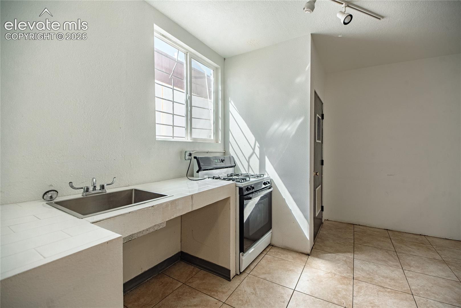 Stainless steel sink, white wall finishes, track lighting, tiled flooring, and a window with security bars