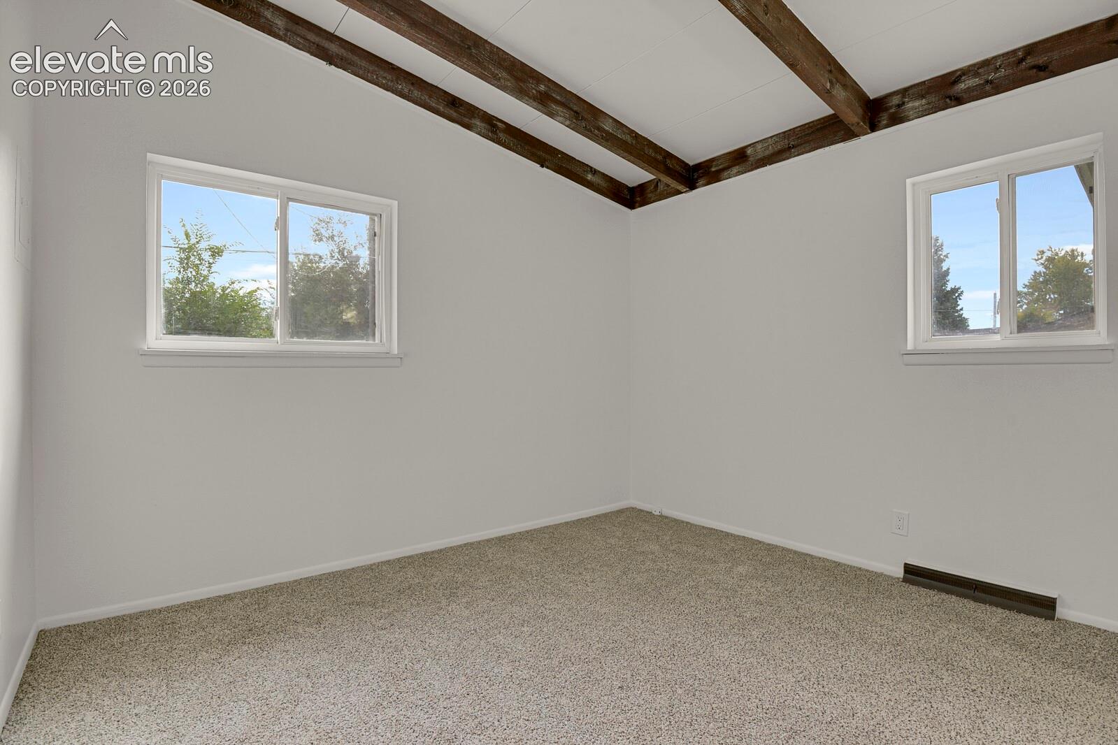 Vaulted ceiling featuring exposed wood-finish beams, neutral carpeting, and dual sliding windows