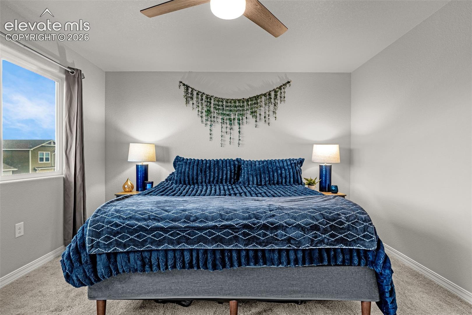 Spacious bedroom with light gray walls, plush carpeting, a large window, a ceiling fan with integrated lighting, and white baseboards