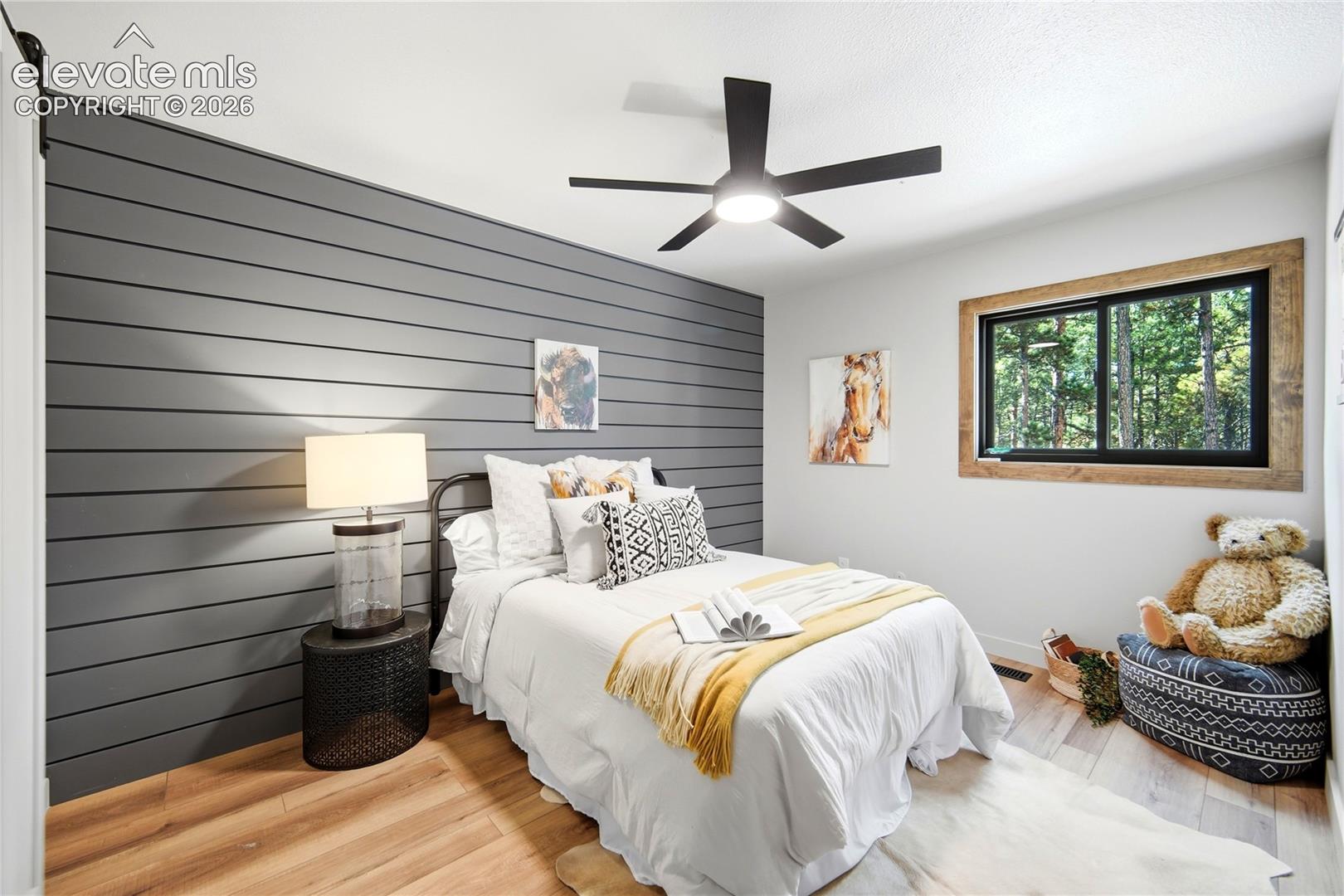 Gray shiplap accent wall, wood-finish flooring, sliding window with rustic timber casing, and modern matte black ceiling fan with integrated light