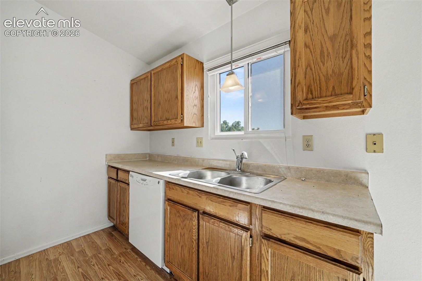 Kitchen featuring wood cabinetry, integrated solid-surface countertop, stainless steel double-basin sink with faucet, white dishwasher, and wood-finish flooring