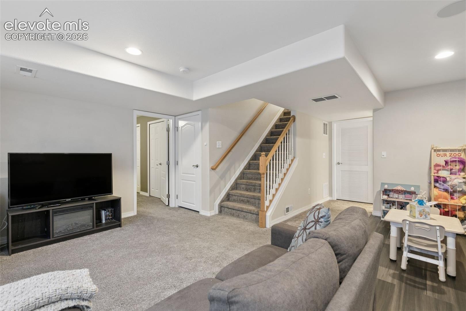 Spacious room featuring neutral carpeting, recessed lighting, white interior doors, and a staircase with a wood handrail