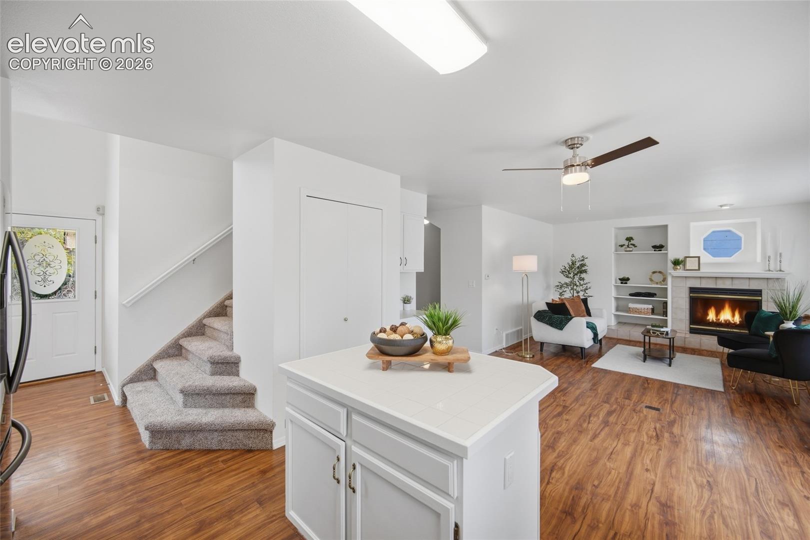 Interior featuring wood-finish flooring, a kitchen island with tile countertops, a fireplace with a masonry surround, built-in shelving, and a ceiling fan