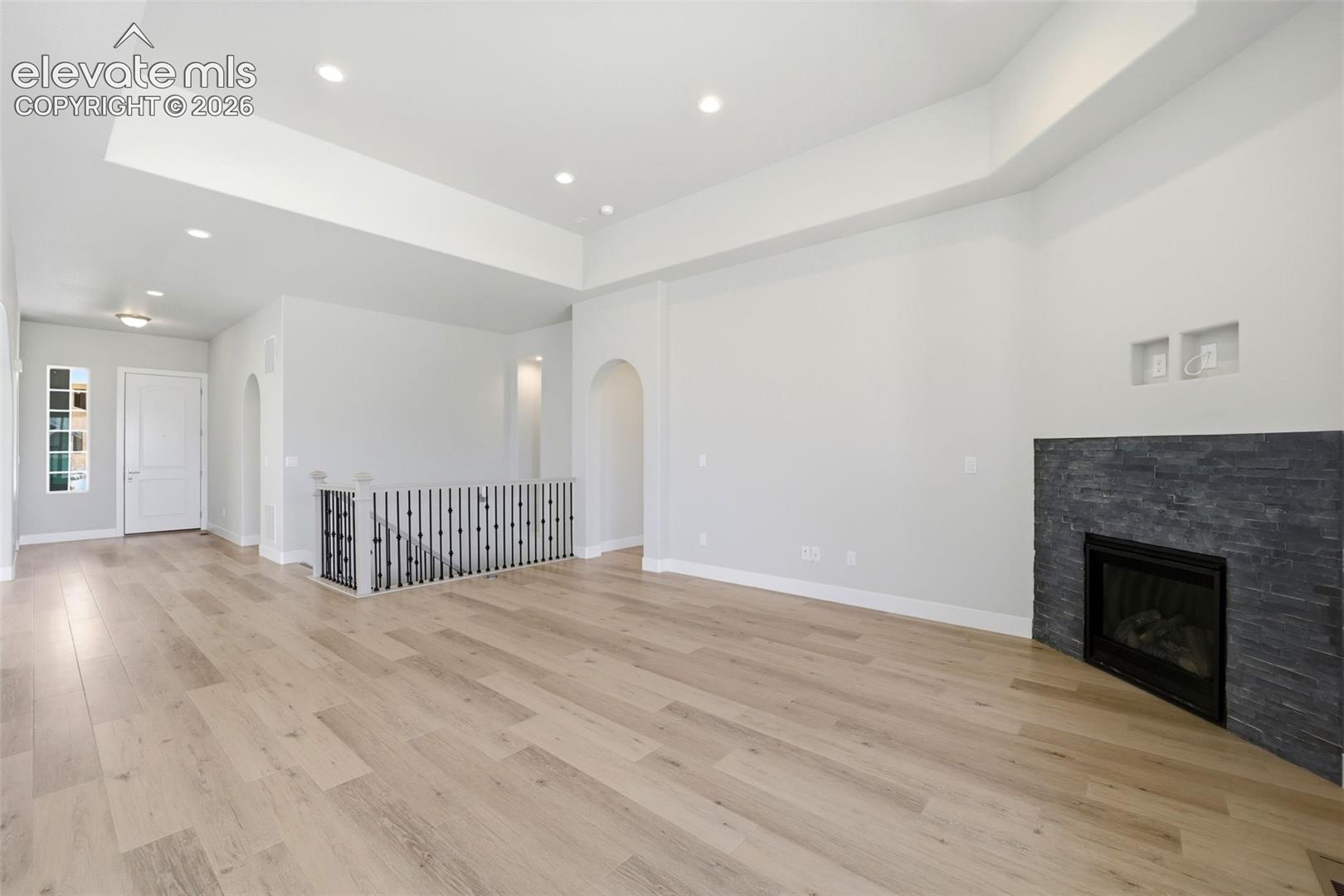 Open-concept room featuring a tiered tray ceiling, wood-finish flooring, stone-tiled fireplace, recessed lighting, and a metal balustrade