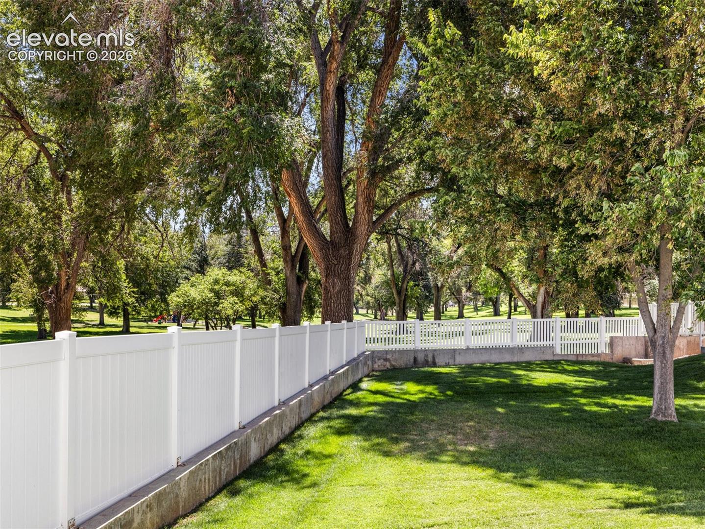 Expansive grassy area with mature trees, featuring a white privacy fence and a white picket fence on a concrete retaining wall