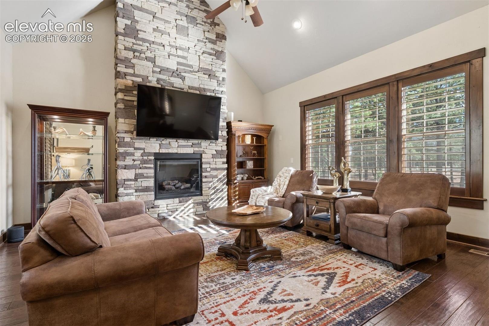 Vaulted ceiling with a central fan, floor-to-ceiling stone fireplace, and wood-finish flooring, accented by large windows with dark wood trim