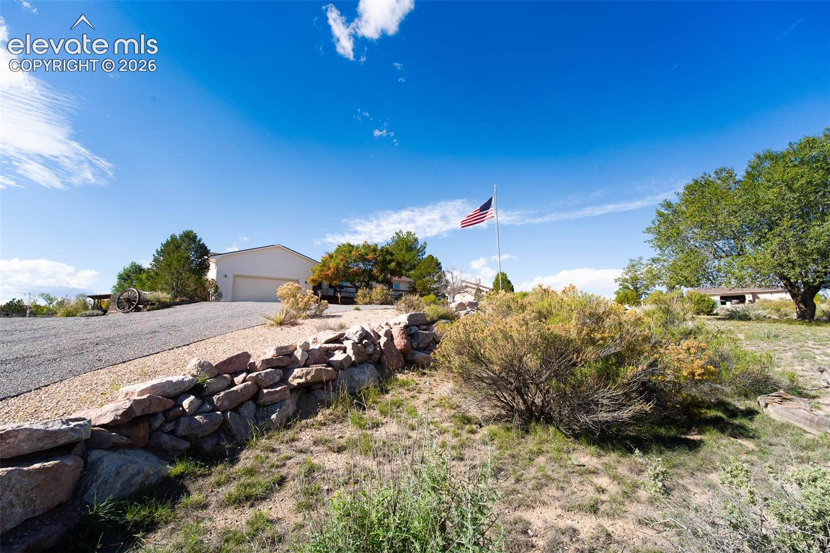 Exterior featuring a stone retaining wall, gravel driveway, attached garage, and landscaping with mature trees